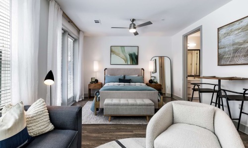 A view of a bed, sofa, and dining space with large windows and ample lighting at The Met apartments. 
